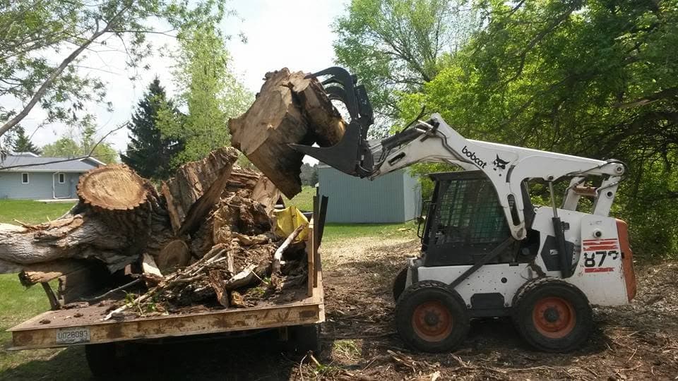 A Bobcat skid-steer loads a large stump section onto a flatbed trailer, metal building behind